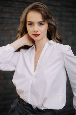 Kendine güvenen bir iş kadınının portresi boynuna dokunuyor, şık bir beyaz gömlek ve koyu renk pantolon giyiyor, profesyonellik saçıyor ve dengede duruyor.