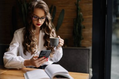 Bardaklı iş kadını kahve içiyor ve kafede uzaktan çalışırken akıllı telefon kullanıyor.