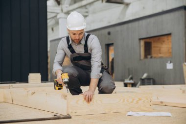 Carpenter using cordless screwdriver assembling wooden frame for modular building in prefabrication workshop clipart