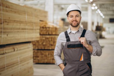 Marangoz tulum giyiyor ve kasketli gülümsüyor ve kereste deposunda ahşap üretimini denetliyor. Modüler yapı endüstrisinde.