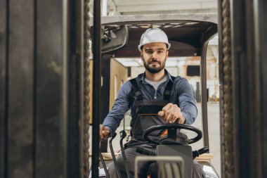 Güvenlik kaskı takmış bir forklift operatörü yoğun bir fabrikadan ustaca manevra yapıyor. Üretimde makine entegrasyonu gösteriliyor. Hassasiyet ve beceri bu dinamik inşaat kariyerinde hayati önem taşıyor.