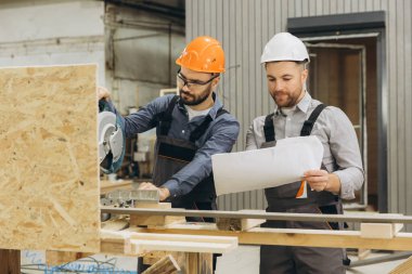 İnşaat işçileri elektrikli testereyle modern fabrikada prefabrik ev inşa ediyorlar ve planları kontrol ediyorlar.