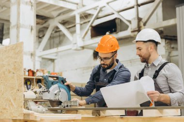 Atölyedeki inşaatçılar dairesel testere ve planı hassasiyetle ve takım çalışmasıyla prefabrik bir ev inşa ediyorlar. Endüstriyel ortam üretim atmosferine ekleniyor