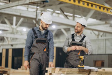 Kask ve tulumlu iki mühendis büyük bir fabrika ortamındaki modüler bir inşaat projesinin planlarını tartışıyor.