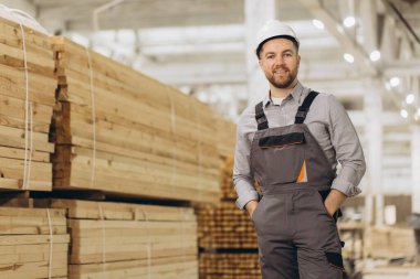 Sıkı şapkalı ve iş tulumlu, elleri ceplerinde gülümseyen, ambardaki kereste yığınlarının önünde duran bir yönetici.