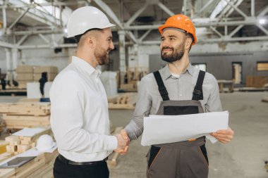 Mimar ve inşaat işçisi modüler bir inşaat fabrikasında el sıkışıyor, planları tartışıyor ve bir proje üzerinde işbirliği yapıyor.