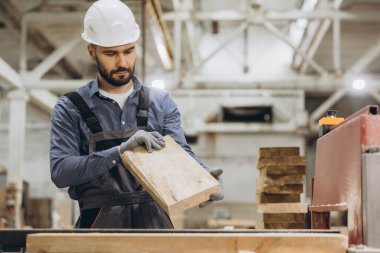 Marangoz koruyucu giysiler giyerek bir marangoz ahşap fabrikasında ahşap bir kalası dikkatle incelerek modüler yapım kalitesini sağlama alıyor.