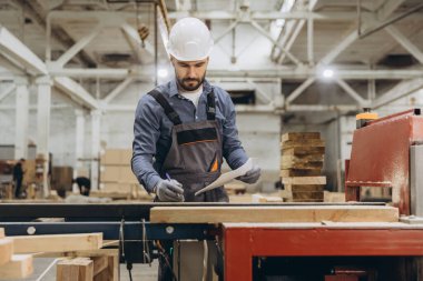 Marangoz güvenlik kaskı takıyor ve iş tulumu giyiyor. Tahtaya not alıyor. Tahta işleme fabrikasındaki tahtaları inceliyor.