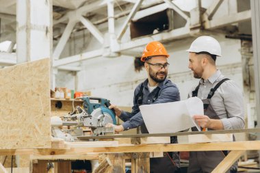 İki mühendis, bir atölyede odun kesmek için dairesel testere kullanırken takım çalışması ve hassasiyetini gösteren planları tartışıyorlar.