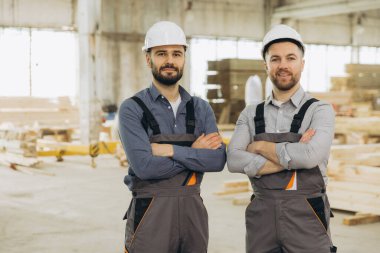 İki kendine güvenen inşaat işçisi, modüler yapı fabrikasında çapraz silahlarla, prefabrik inşaatlarda takım çalışması ve uzmanlık sergileyerek gururla duruyorlar.