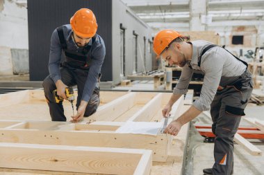 Bir fabrikada modüler yapı için kablosuz tornavida ve mezura kullanarak prefabrik ahşap paneller monte eden iki inşaatçı.