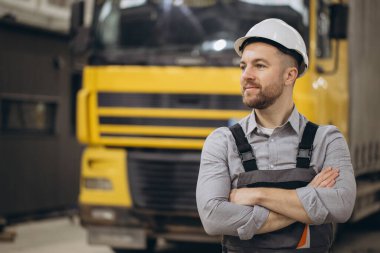 Modüler yapı fabrikasında, kamyonun önünde kolları çapraz duran kasklı ve tulumlu kendinden emin mühendis.