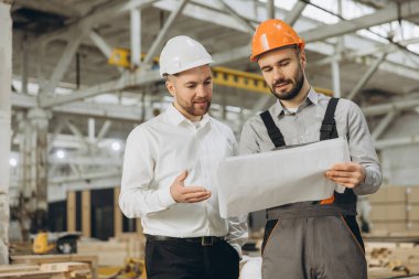 İki mühendis, araçlar ve teknolojiyle çevrili bir inşaat projesine odaklanmış modern bir fabrikanın planlarını incelerken işbirliği yapıyor ve tartışıyorlar.