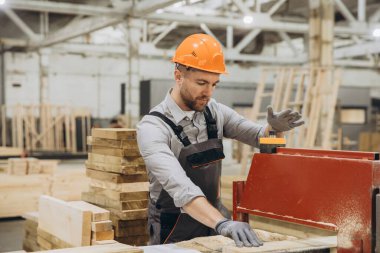 Marangoz koruyucu kask ve eldiven takıyor. Kereste işleme fabrikasında ahşap planlama makinesi kullanıyor. Modüler üretim için.