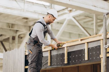 Güvenlik kaskı takan inşaat işçisi, fabrikada prefabrik evin ahşap yapısını birleştiren elektrikli tornavida kullanıyor.