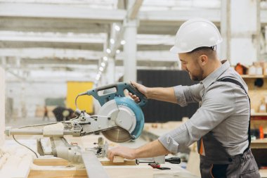 Marangoz kaskını dairesel testere kullanarak takıyor prefabrik bir ev fabrikasında ahşap kalas kesiyor.