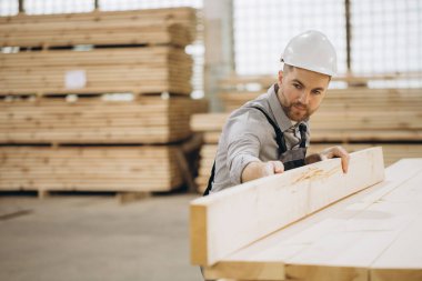 Marangoz kereste deposundaki ahşap tahtaları kontrol ediyor. Modüler bir inşaat projesi üzerinde çalışıyor. Kaliteyi ve hassasiyeti sağlıyor.