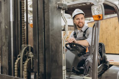 Gülümseyen depo işçisi fabrikada forklift kullanıyor, modüler inşaat için kaldırma ekipmanları kullanıyor.