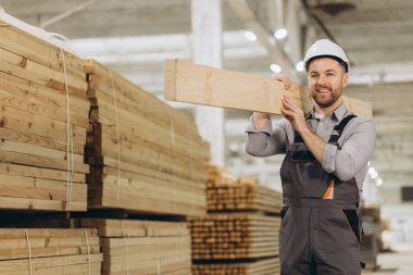 Gülümseyen marangoz büyük kereste deposunda ahşap kalas taşıyor. İnşaat ve imalat için kaliteli kereste sergiliyor.