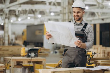 Gülümseyen inşaat işçisi prefabrik bir ev fabrikasının planlarını inceliyor, modüler inşaatta kalite ve hassasiyet sağlıyor.