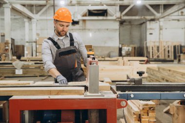 Marangoz koruyucu elbiseler giyiyor. Atölyede testere kullanarak tahta kesiyor. Modüler bina inşaatı için parçalar hazırlıyor.
