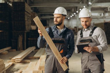 Veri analizi ve kontrol için tablet kullanan iki mühendis kereste fabrikasındaki ahşap kalasları inceliyor.