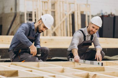 İki inşaatçı, marangozluk atölyesinde prefabrik ev iskeleti montajı için matkap ve ruh seviyesini kullanıyor.