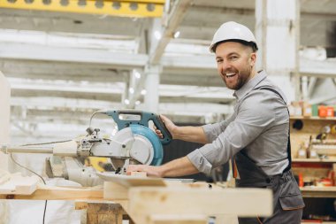 Gülümseyen marangoz dairesel testereyle ahşap kalas kesiyor, güvenlik kaskı takıyor ve iş tulumu giyiyor, açık ve havadar bir atölye ortamında
