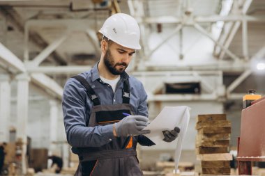 Güvenlik kaskı takan ve iş tulumu giyen inşaat işçisi planları inceliyor ve prefabrik bir ev fabrikasında notlar alıyor.