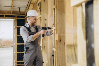 İnşaat işçisi modern modüler ev inşaatı için kablosuz tornavida kullanarak inşaat alanında prefabrik ahşap paneller kuruyor.