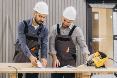 Sert şapka ve tulum giyen iki müteahhit planları inceliyor ve fabrikadaki tezgahın üzerinde testere hazırlıyor.