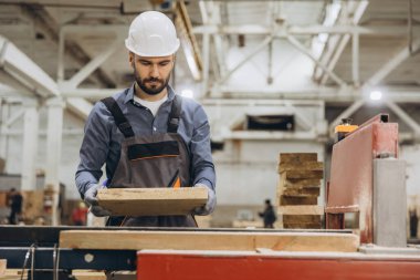 Marangoz güvenlik kaskı takıyor kereste fabrikasında ahşap tahtaları inceliyor, prefabrik ev projesi üzerinde çalışıyor.