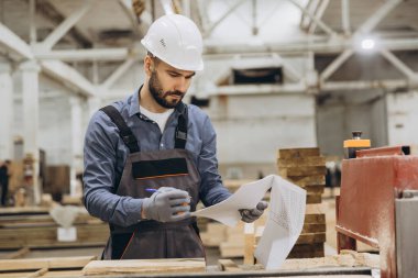 Marangoz koruyucu iş giysisi giyiyor. Modüler bina inşaatı için kereste işleme fabrikasında proje planını kontrol ediyor.