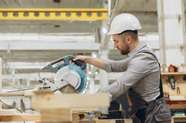 Marangoz güvenlik kaskı takıyor. Dairesel testere kullanıyor. Ahşap işçiliği yapan bir fabrikada modüler yapım için tahta kesiyor.