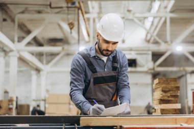 Başlık takan ve iş tulumları giyen yönetici, modüler yapı fabrikasındaki tahta tahtaları incelerken panoya notlar yazıyor.