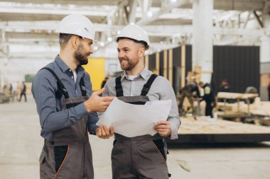 İki mühendis planlarını incelerken aynı zamanda gülümsüyor ve bir modüler yapı fabrikasında proje detaylarını tartışıyor.