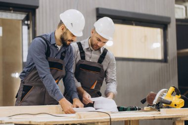 Başlık ve tulum giyen iki müteahhit, bir marangozhane atölyesindeki modüler yapı projesinin planlarını kontrol ediyor.