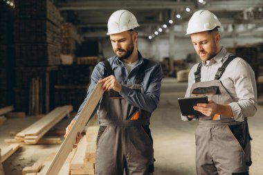 İki inşaat işçisi ahşap bir kalası inceliyor ve modüler inşaat endüstrisi fabrikasında tablet kullanıyorlar.