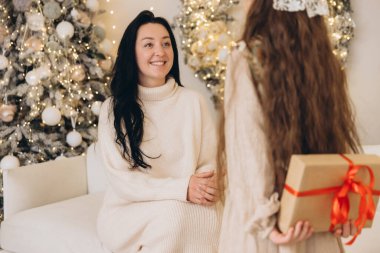 Yılbaşı hediyesini arkasına saklayan bir kız, annesinin süsleyerek Noel ağacının yanında oturuyor, sıcak aile ortamı yaratıyor.