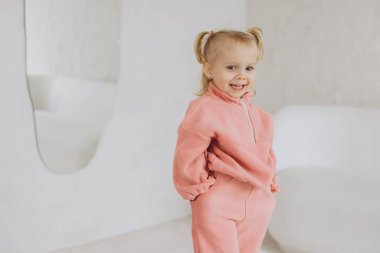 İki at kuyruklu genç kız pembe yün kazak ve pantolon giyiyor, elleri ceplerinde, beyaz odada gülümsüyor.