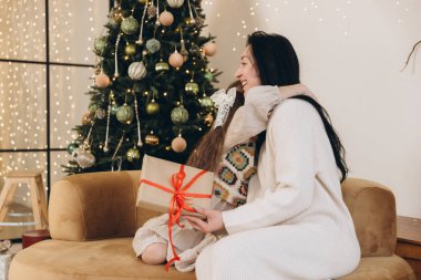 Kızı, mutlu annesine Noel hediyesi veriyor. Süslü ağacın yanındaki kanepede sarılıyor.