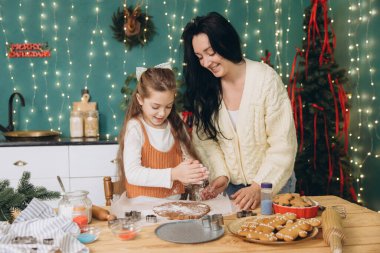 Anne ve kızı birlikte Noel kurabiyesi yapıyorlar. Mutfakta, ışıklarla ve noel ağacıyla süslenmiş.