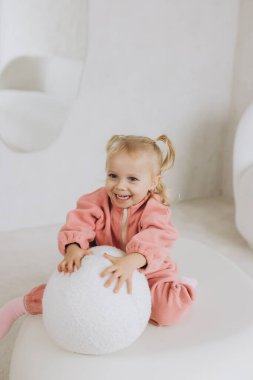 Pembe postu olan küçük kız beyaz modern mobilyaların üzerinde oturuyor, beyaz tüylü topla oynuyor, gülümsüyor ve mutlu görünüyor.