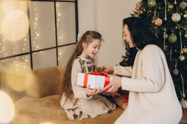 Gülümseyen kız, annesi tarafından hediye edilen Noel hediyesini açıyor, arka planda sıcak ışıklar olan dekore edilmiş ağacın yanındaki koltukta oturuyor.