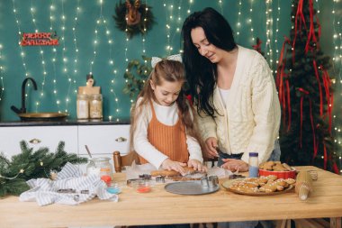 Anne ve kızı, Noel ruhunu kucaklayarak, sıcacık dekore edilmiş bir mutfakta, kurabiye hamurunu neşeyle yuvarlıyorlar.