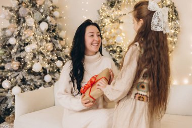 Kızı, gülümseyen annesine Noel hediyesi veriyor. Süslü Noel ağacının yanındaki beyaz koltukta oturuyor.