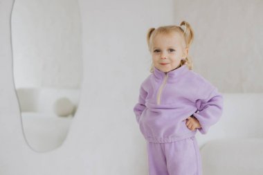 Mor eşofmanlı şirin küçük kız, büyük bir aynayla modern, minimalist bir iç mekanda poz verirken gülümsüyor.