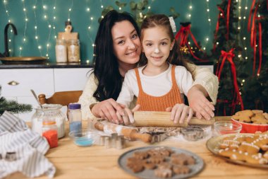 Anne kızına Noel kurabiyesi pişirmeyi öğretiyor. Mutfak masasında ahşap raptiyeyle hamuru yuvarlıyor. Arka planda Noel ağacı var.