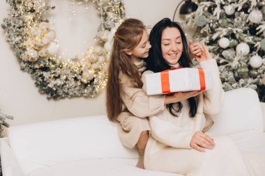 Kız, gülümseyen annesine yılbaşı hediyesi veriyor. Süslü Noel ağacının yanındaki beyaz koltukta oturuyor.