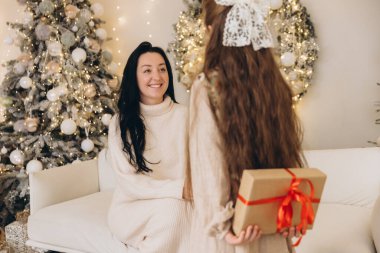 Arkasında Noel hediyesini saklayan bir kız annesine sürpriz yapıp Noel ağacının yanındaki koltukta oturuyor.